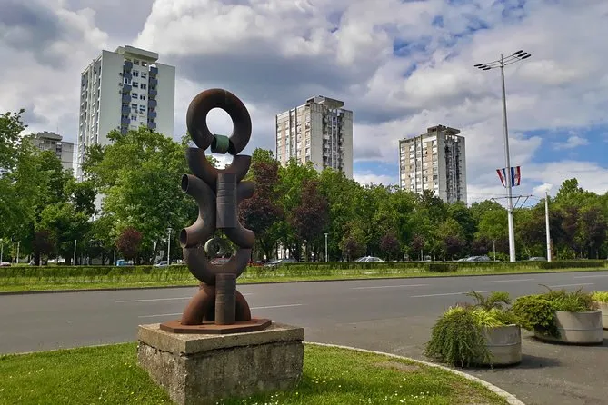An abstract rust-coloured sculpture on a concrete plinth beside a Novi Zagreb boulevard, with socialist-era high-rise blocks behind